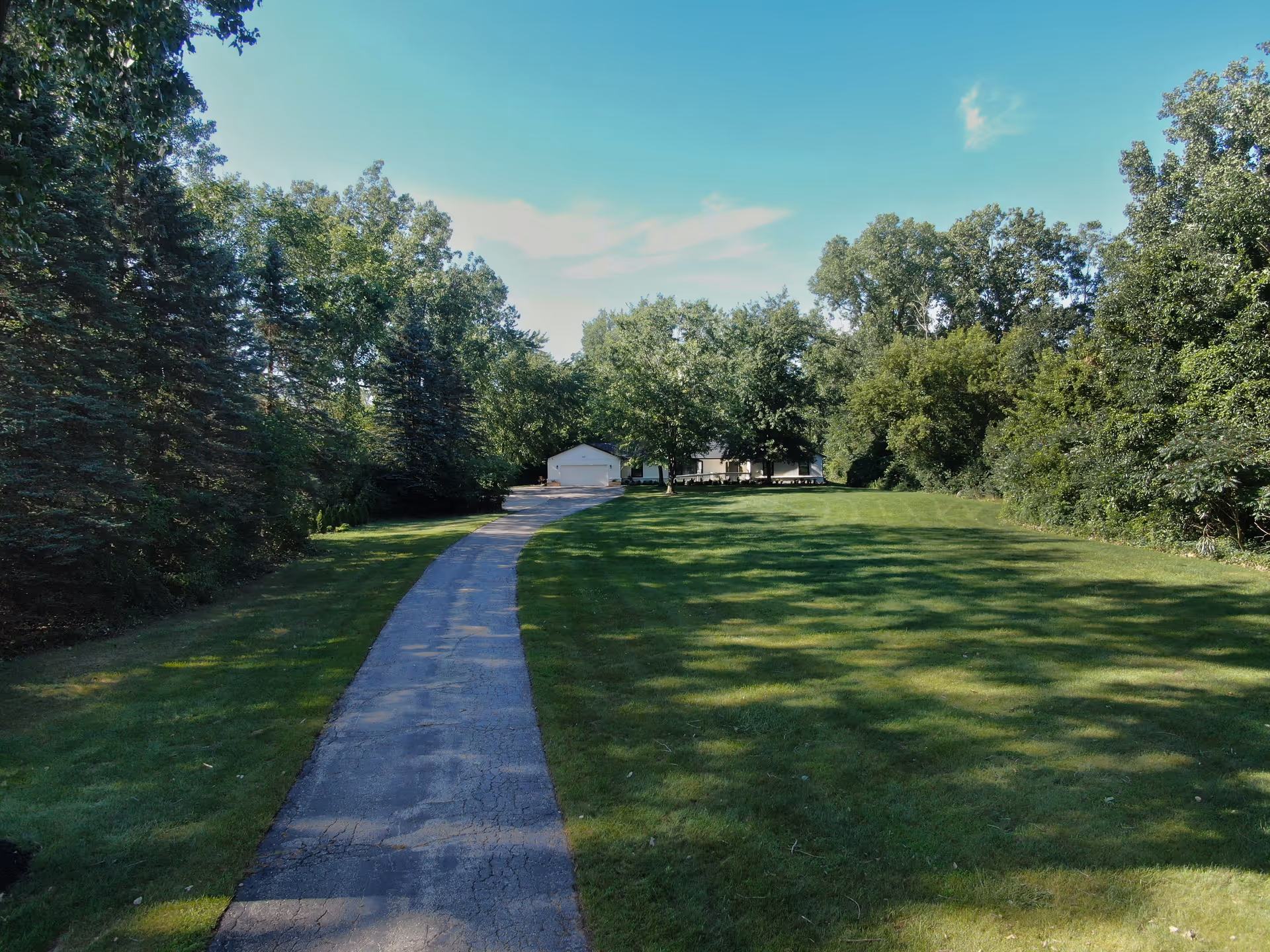 A long paved driveway leading to a white building with a garage, surrounded by green grass and tall trees under a clear blue sky.