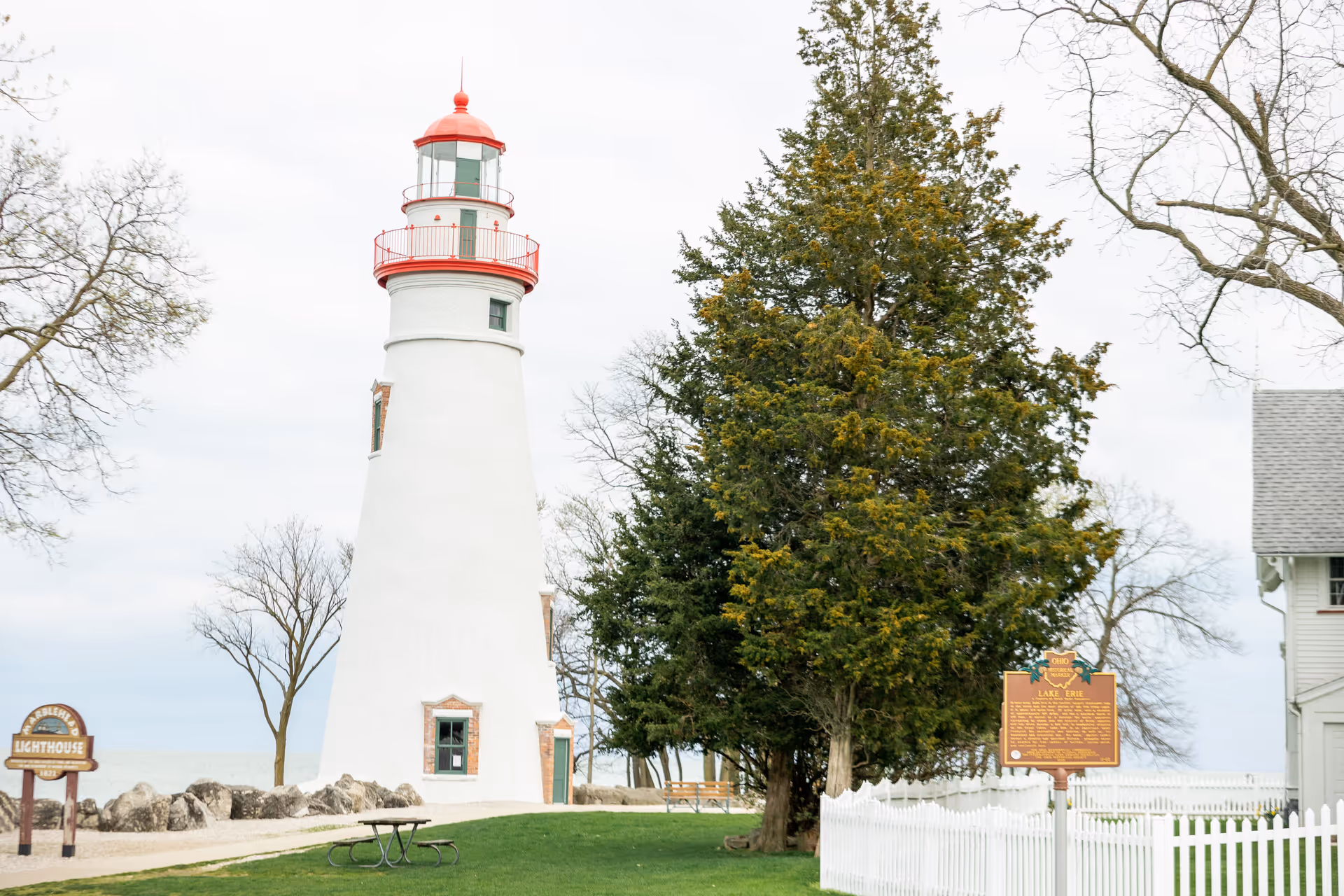 A white lighthouse with a red top and railing stands near a grassy area with a picnic table. There are trees, a white picket fence, and informational signs around the lighthouse. The sky is overcast.