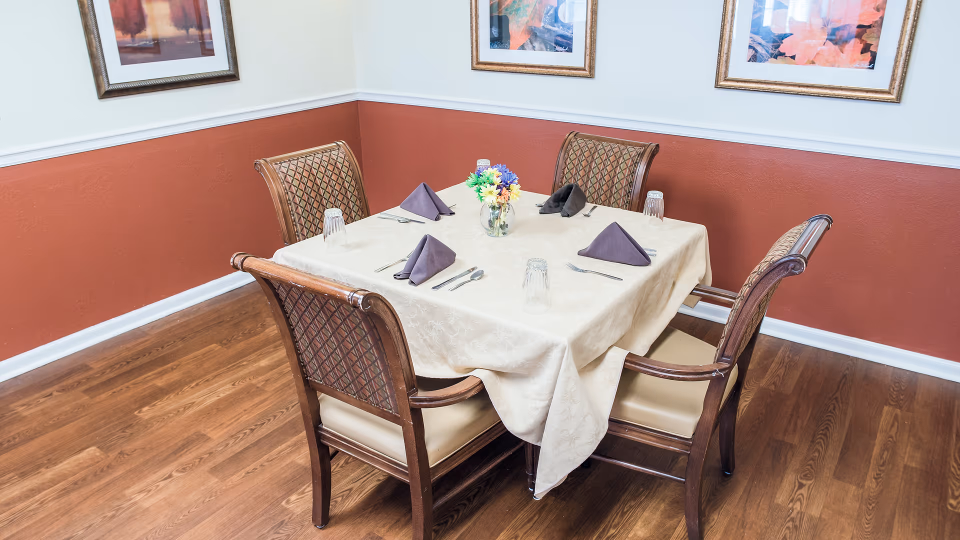 A square dining table set for four with folded napkins, glasses and a small floral centerpiece in a room with framed art and wood flooring.