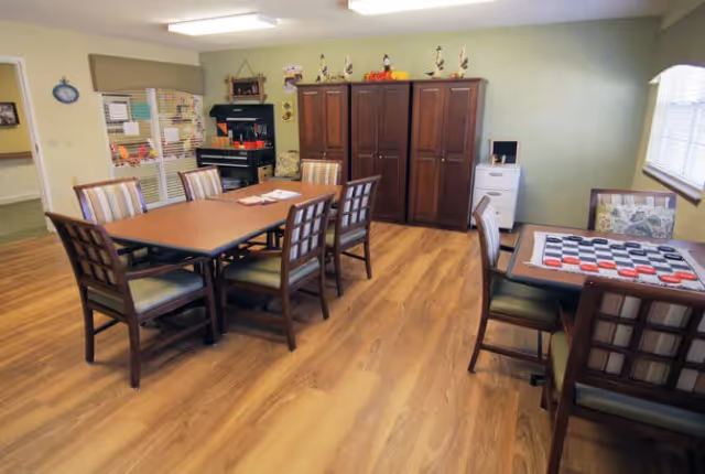 A senior living community activity/dining room with multiple tables and chairs, a checkerboard game on one table, and storage cabinets against a green wall.
