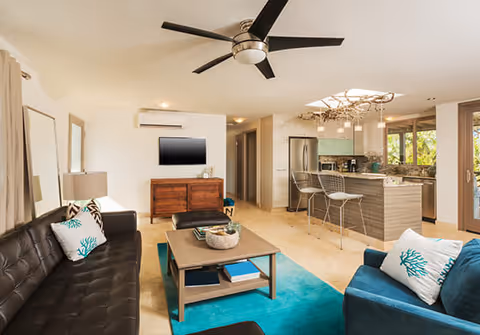A modern open-concept living room and kitchen area featuring a black leather sofa with white and teal coral-patterned pillows, a blue armchair with a matching pillow, a wooden coffee table on a teal rug, a ceiling fan with black blades, a wall-mounted flat-screen TV above a wooden cabinet, and a kitchen with a breakfast bar, two metal bar stools, stainless steel refrigerator, and large windows letting in natural light.