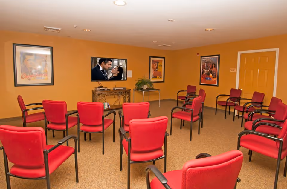 A room with multiple red cushioned chairs arranged in rows facing a wall-mounted television screen displaying a scene from a movie. The walls are painted orange and decorated with framed movie posters. There is a small table with a plant and some electronic devices under the TV.