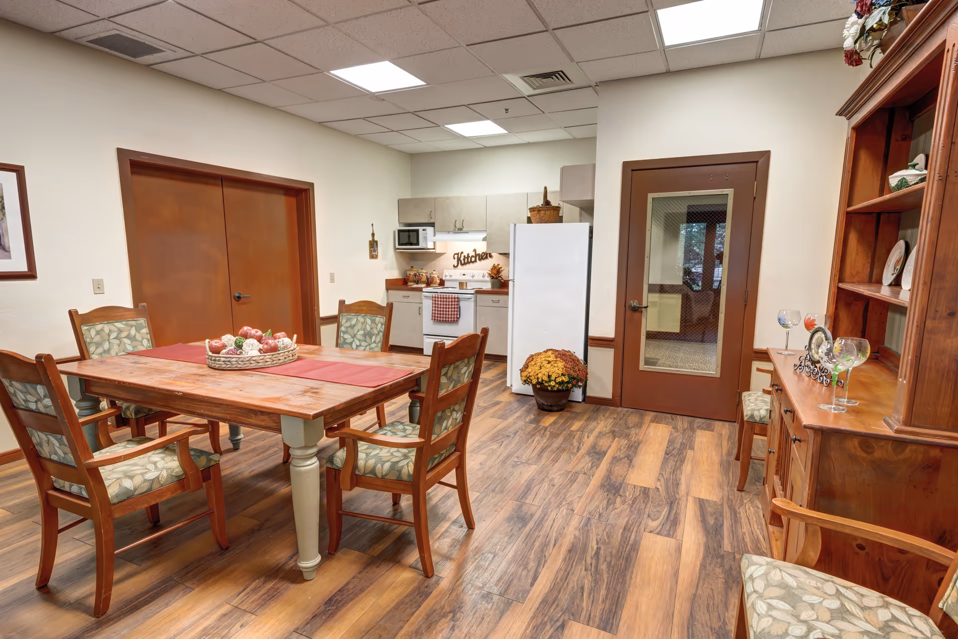 A cozy dining area with a wooden table and four cushioned chairs featuring a leaf pattern. The table has a red runner and a decorative basket with artificial fruit. In the background, there is a small kitchen with a white refrigerator, stove, microwave, and cabinets. A wooden hutch with decorative plates and glasses is on the right side. The room has wood-look flooring, beige walls, and a ceiling with fluorescent lights.