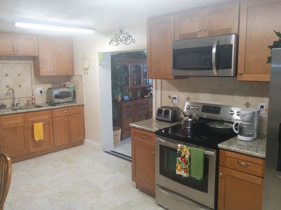 A kitchen with wooden cabinets, granite countertops, and tiled backsplash. The kitchen features a stainless steel microwave above a stainless steel stove with a pot on it, a toaster, and a white coffee maker. There is a yellow towel hanging on a cabinet near the sink and two dish towels hanging on the oven door. The floor is tiled, and there is an open doorway leading to another room with wooden furniture and plants.