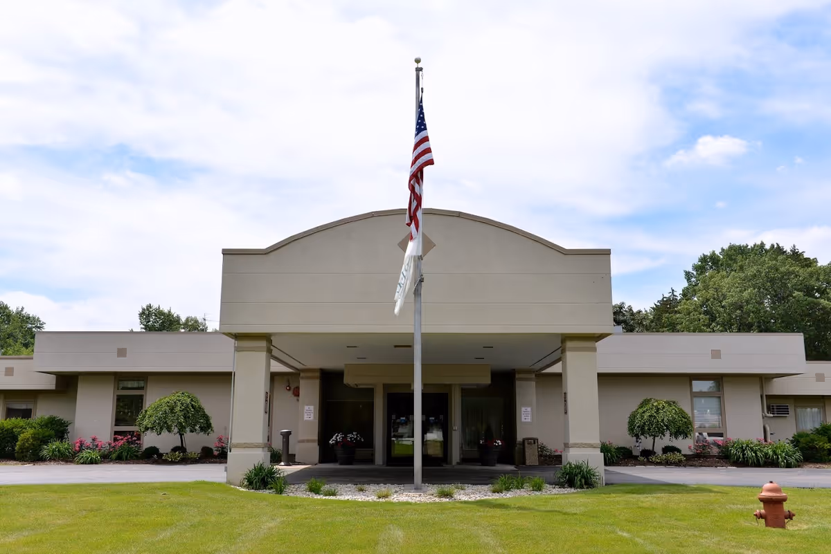 Front exterior view of a single-story beige building with a covered entrance, an American flag on a flagpole in front, manicured bushes and flowers along the building, and a fire hydrant on the grass lawn.