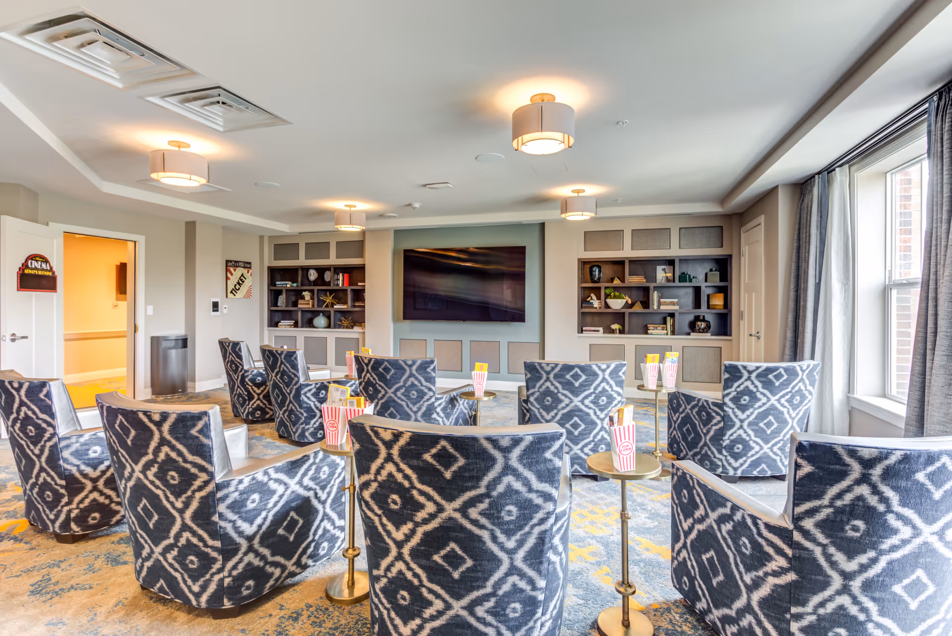 A cozy media room with patterned armchairs arranged in rows facing a large flat-screen TV mounted on the wall. Small round tables between chairs hold popcorn containers. The room has built-in shelves with decorative items and books, soft lighting from ceiling fixtures, and large windows with curtains letting in natural light.