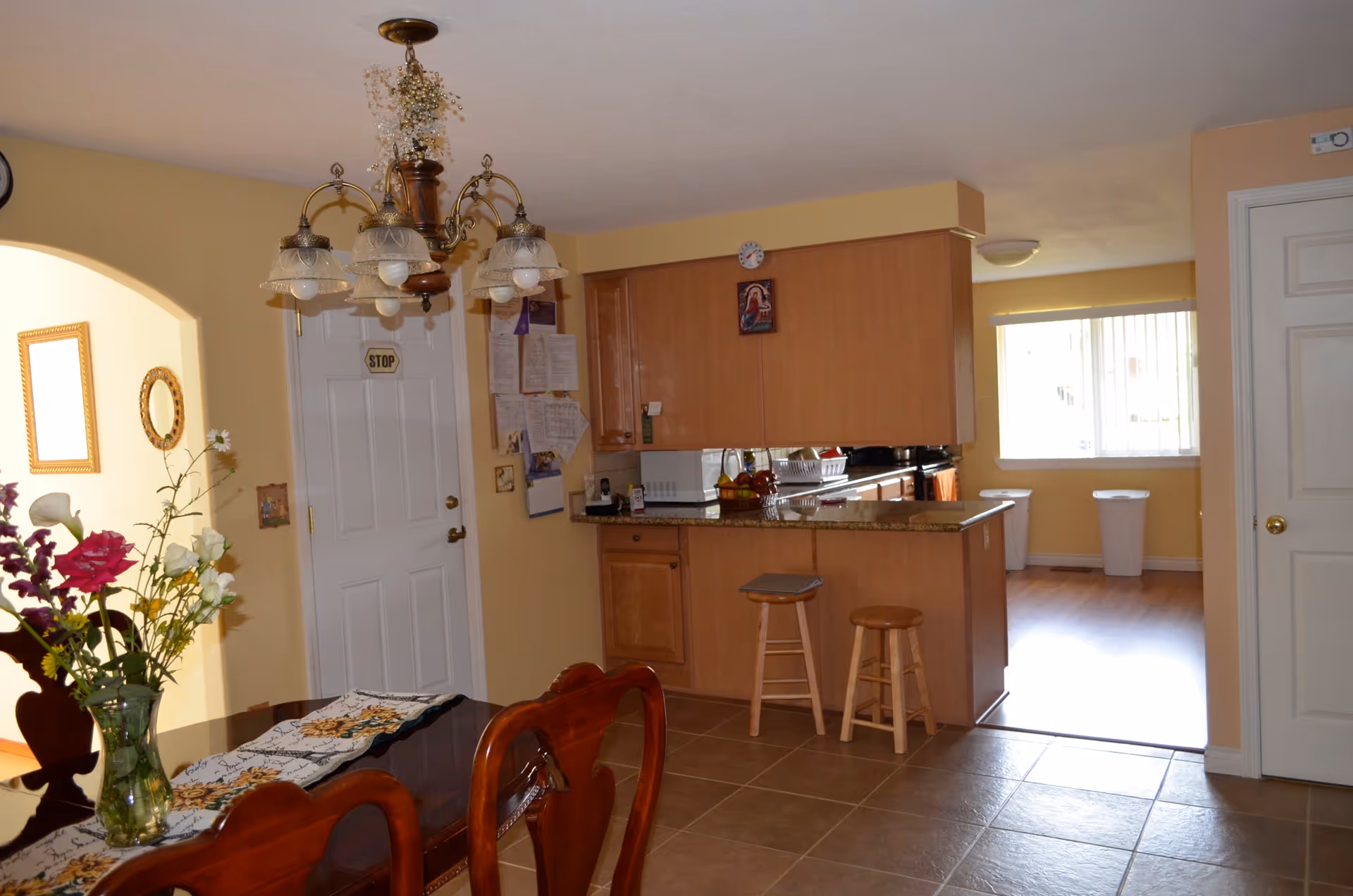 Interior view of a dining area and kitchen in a residential facility. The dining area has a wooden table with a floral table runner and a vase of flowers. Above the table hangs a decorative chandelier. The kitchen features wooden cabinets, a granite countertop with two wooden stools, a microwave, and a fruit basket. There is a door with a 'STOP' sign, a bulletin board with papers, and a window with vertical blinds letting in natural light.