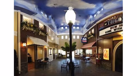 Interior view of a spacious common area in a senior living facility with a high ceiling painted to resemble a sky with clouds. The area features a central lamppost, several benches, tables with chairs, and storefront-style facades including a cafe and a theater marquee. The space is well-lit with natural light coming through high windows.