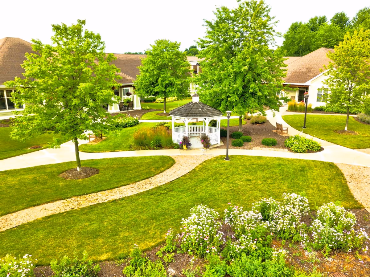 A well-maintained outdoor garden area at The Auberge at Brookfield featuring a white gazebo surrounded by green trees, manicured lawns, flower beds, walking paths, benches, and lamp posts, with buildings visible in the background.