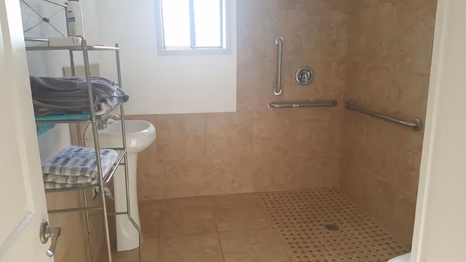 A bathroom with beige tiled walls and floor, featuring a walk-in shower area with grab bars on the walls. There is a small white pedestal sink under a window, and a metal shelving unit holding folded towels and other items.