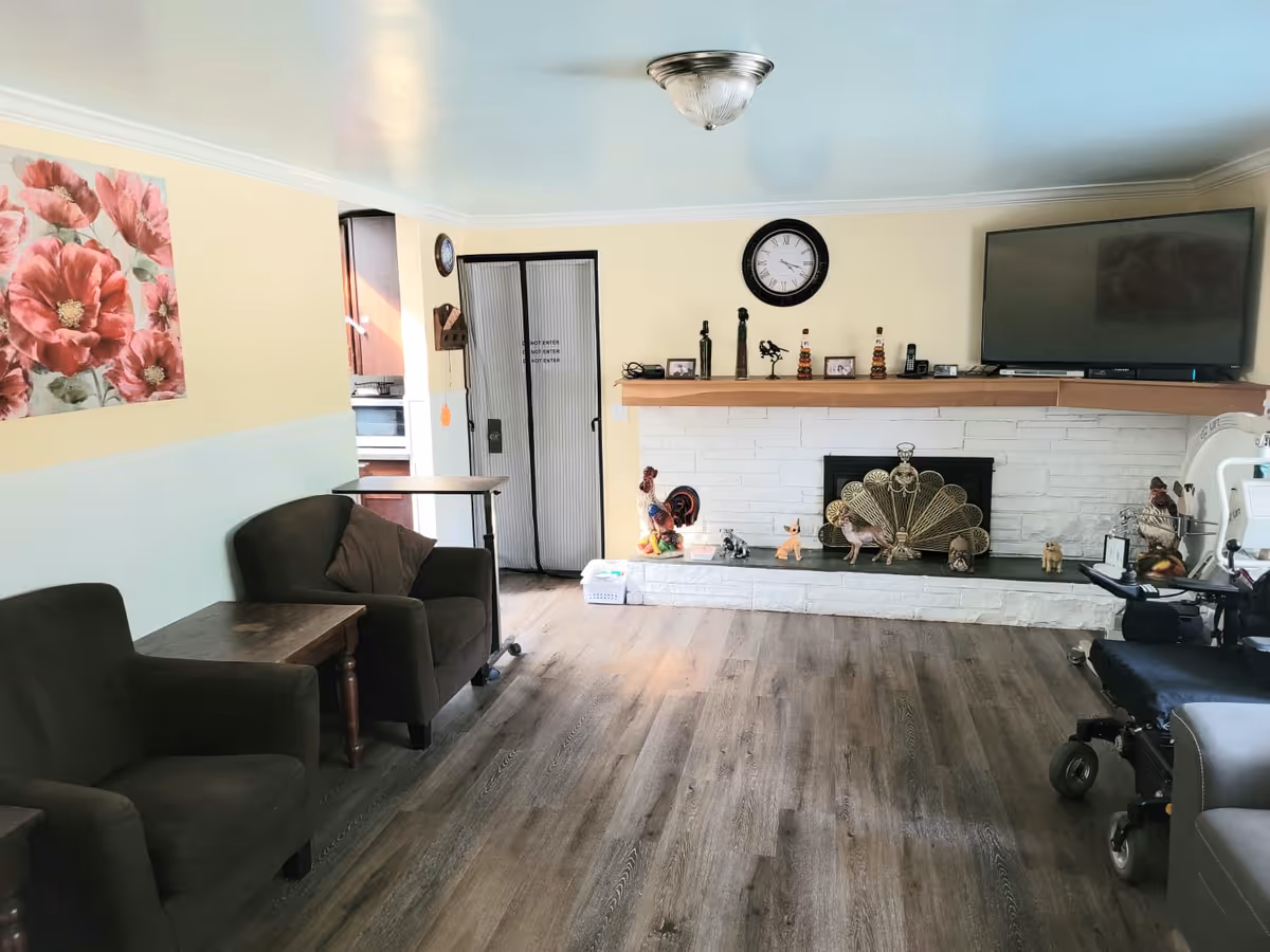 Bright living room with armchairs, a fireplace mantel with decorations, a wall-mounted TV, and wood-look flooring.