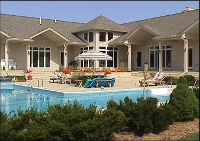 Outdoor view of a senior living facility with a swimming pool in the foreground, surrounded by lounge chairs and umbrellas. The building has large windows and a sloped roof, with greenery and bushes around the pool area.