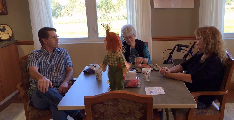 Three people sit around a table in a light-filled communal dining area with a scarecrow centerpiece and a walker by the window.