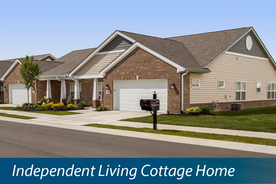 Exterior view of a single-story independent living cottage home with a brick and siding facade, attached garage, mailbox, small front porch, and well-maintained lawn and landscaping under a clear blue sky.