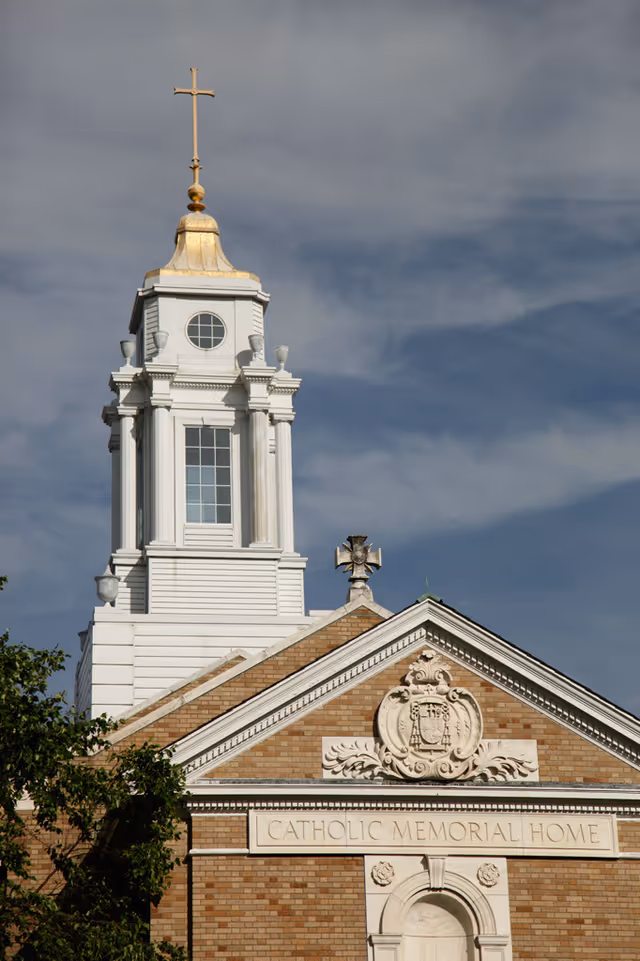 Front facade of the Catholic Memorial Home building with a decorative pediment and a white steeple topped by a gold cross against a cloudy sky.