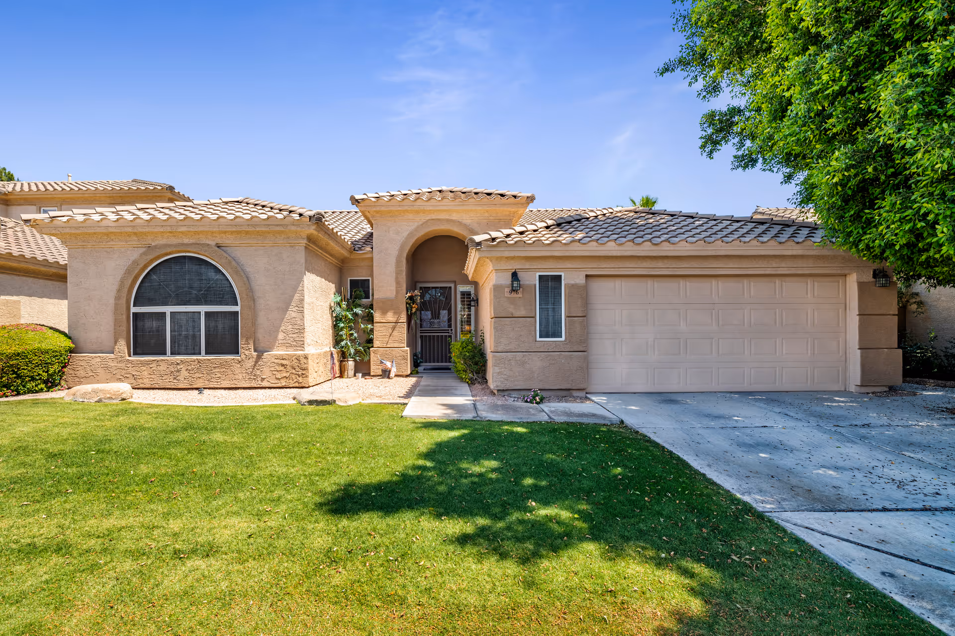 Front exterior view of a single-story residential building with a tiled roof, an arched window, a two-car garage, a small front porch with plants, and a well-maintained green lawn under a clear blue sky.
