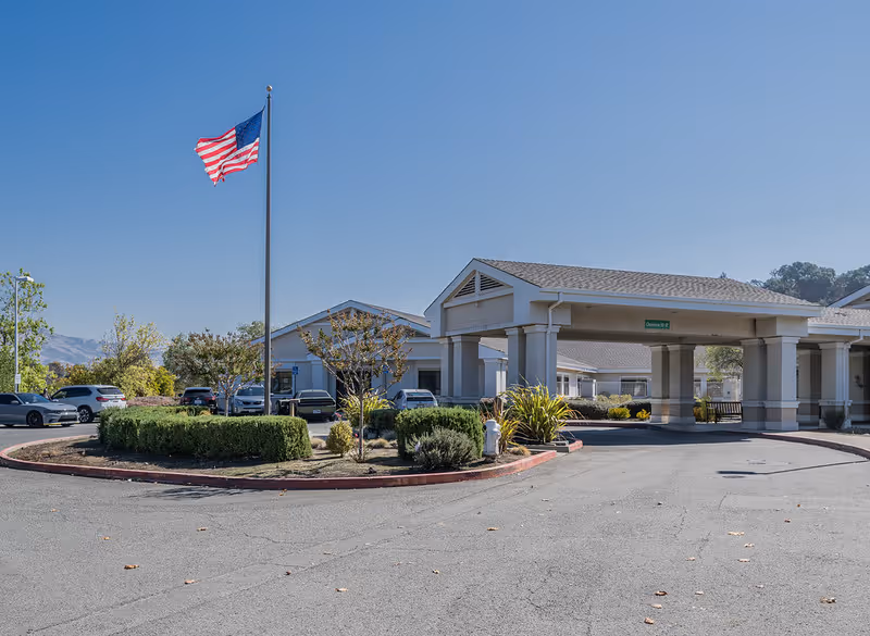 Exterior view of a single-story building with a covered entrance and an American flag on a flagpole in front. Several cars are parked around the building, and there are landscaped bushes and trees. The sky is clear and blue.