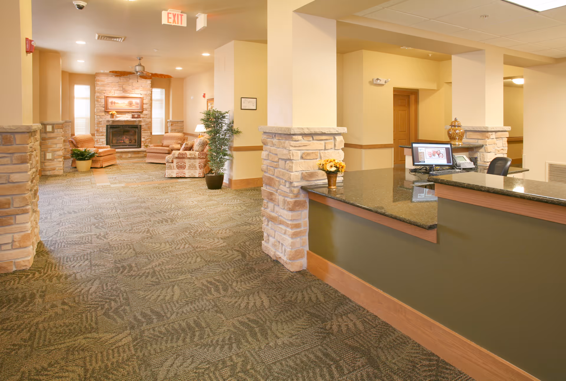 Interior view of a senior living facility lobby area with a reception desk on the right and a seating area with armchairs and a fireplace in the background. The space features stone pillars, patterned carpet, and warm lighting.
