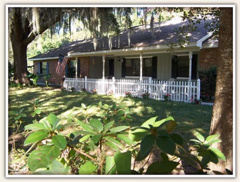 Single-story brick house with a white picket fence, front porch, and an American flag surrounded by trees and shrubs.