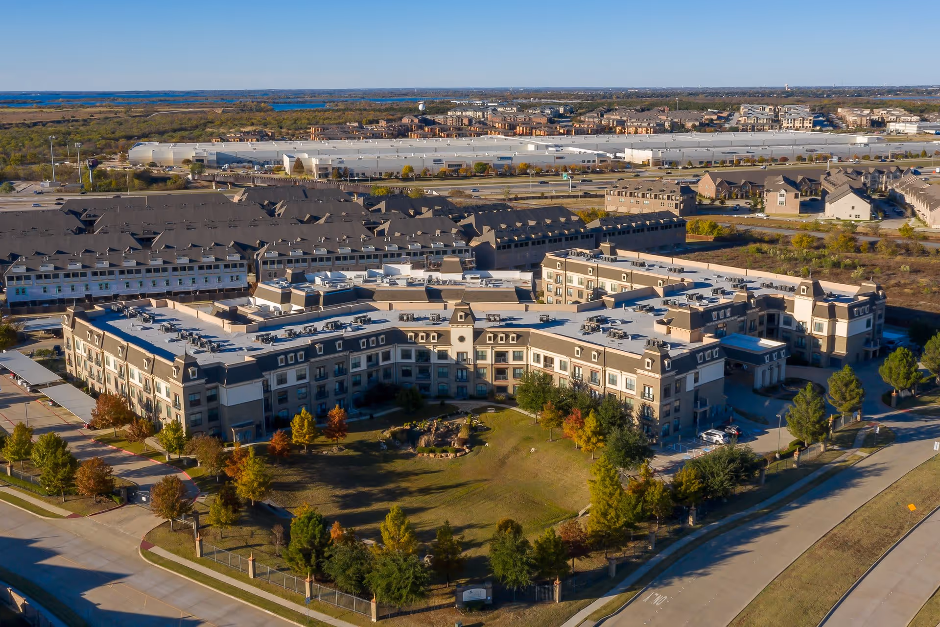 Aerial view of a large senior living facility named Discovery Village At Castle Hills surrounded by landscaped greenery, trees, and roads, with residential buildings and open land in the background under a clear blue sky.