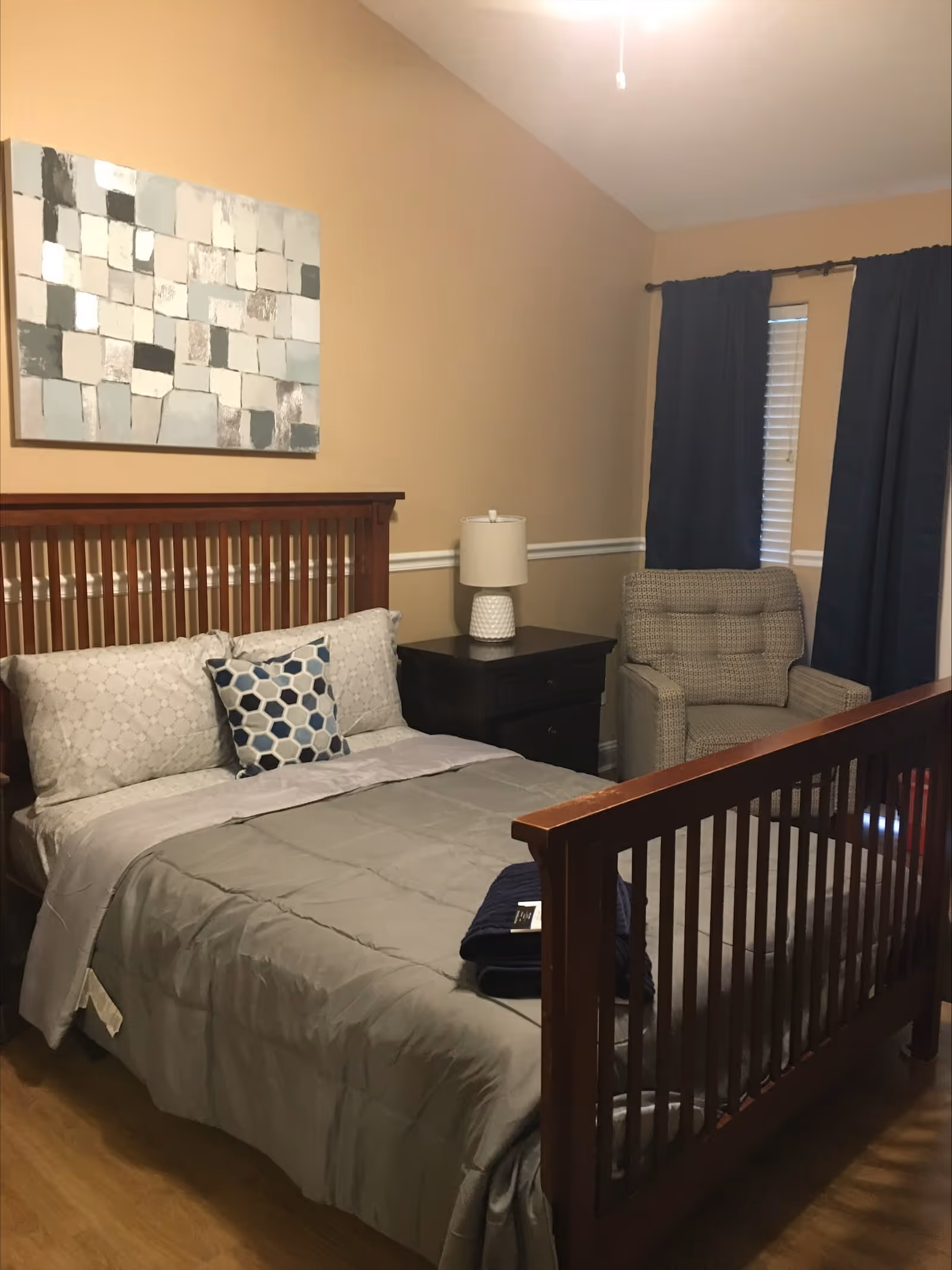 A bedroom with a wooden bed frame, gray bedding, and a decorative pillow with a hexagonal pattern. There is a dark wooden nightstand with a white lamp on it next to the bed. A cushioned armchair is positioned near a window with dark blue curtains. A piece of abstract art with various shades of gray and white hangs on the beige wall above the bed.