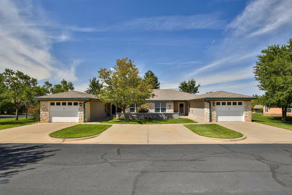 Exterior view of a single-story senior living facility building with two attached garages, a tree in the center, green lawns, and a clear blue sky with some clouds.