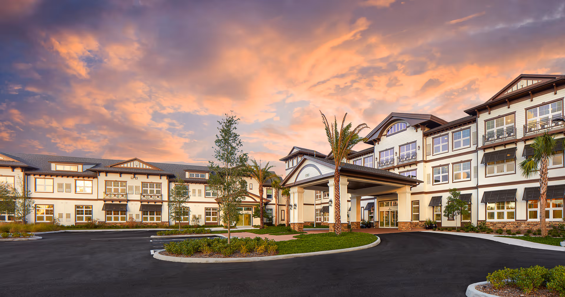 Exterior view of a large, modern senior living facility building with multiple windows, palm trees, and a covered entrance under a colorful sunset sky.