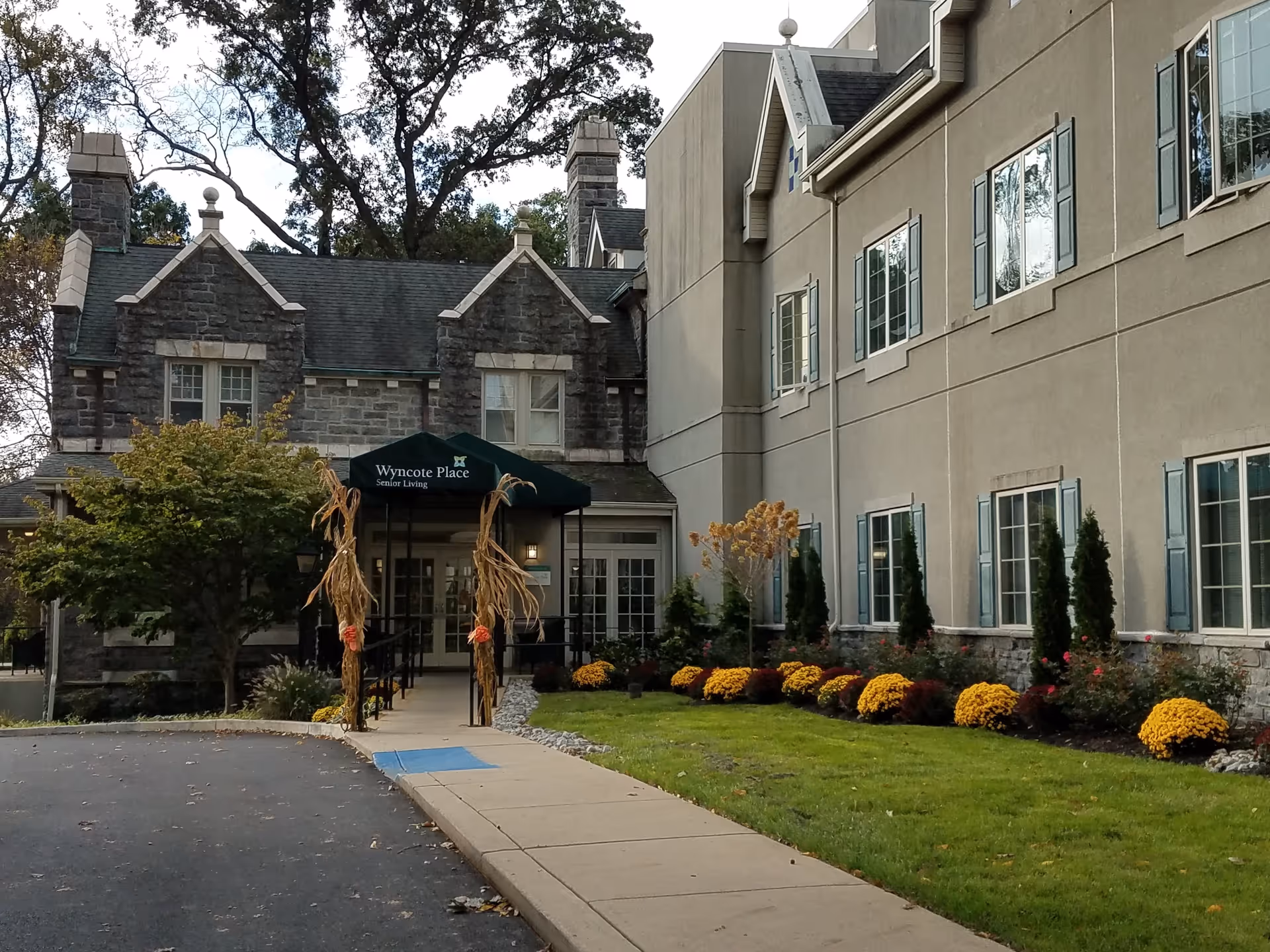 Exterior view of Wyncote Place senior living facility showing a stone and beige building with multiple windows, a green awning over the entrance with the facility name, a paved driveway, sidewalk, and landscaped garden with yellow flowers and small trees.