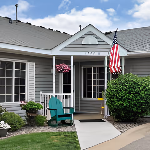 Exterior view of a single-story gray building with white trim, featuring a small covered porch with a green Adirondack chair, an American flag hanging near the entrance, and well-maintained bushes and lawn under a partly cloudy sky.