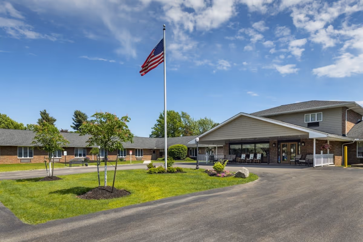 Exterior view of Elderwood Village at Bassett Park facility on a sunny day, showing a single-story building with a covered entrance, an American flag on a flagpole, green lawn with small trees and shrubs, and a clear blue sky with some clouds.