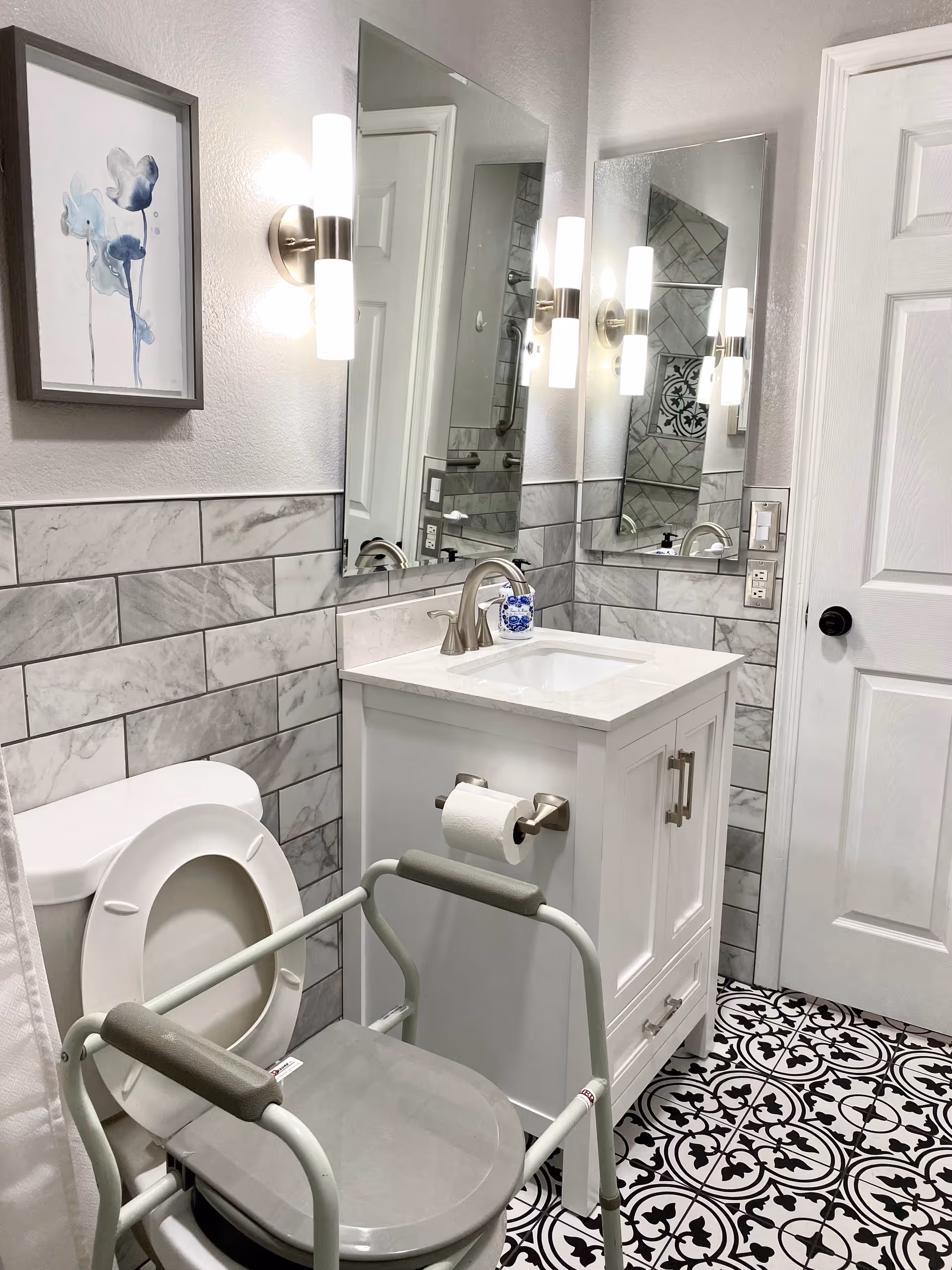 A modern bathroom with a white toilet equipped with a gray raised toilet seat and support frame. The bathroom features a white vanity with a sink, a silver faucet, and a mounted toilet paper holder. The walls are partially tiled with gray marble tiles, and there are two vertical wall lights on either side of a large mirror above the sink. The floor has black and white patterned tiles, and a white door is visible in the background. A framed abstract blue floral painting hangs on the wall above the toilet.