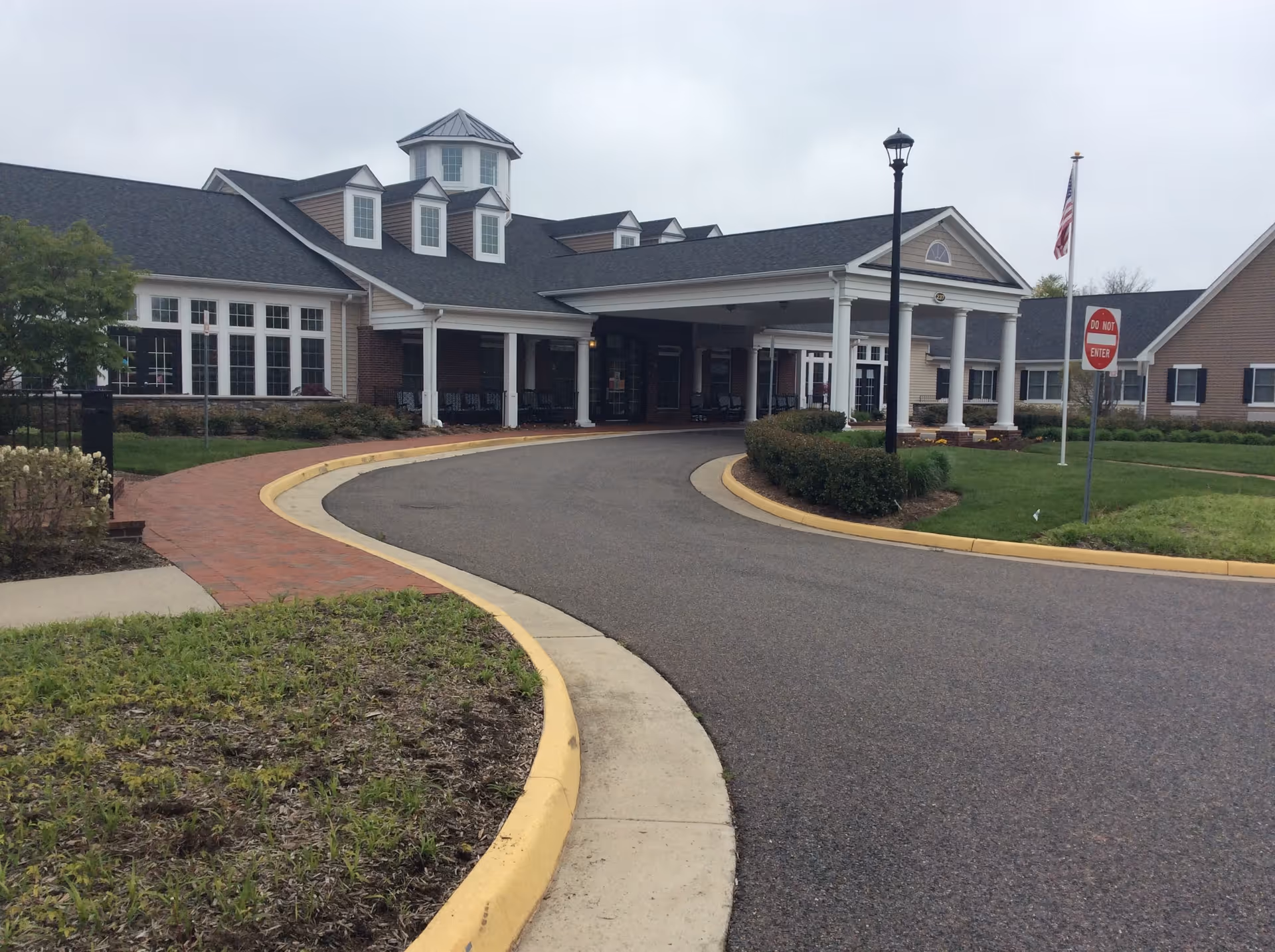 Exterior view of Spring Arbor of Leesburg senior living facility showing the main entrance with a covered driveway, a curved road leading to the entrance, landscaped bushes and grass, a lamppost, an American flag, and a 'Do Not Enter' sign.