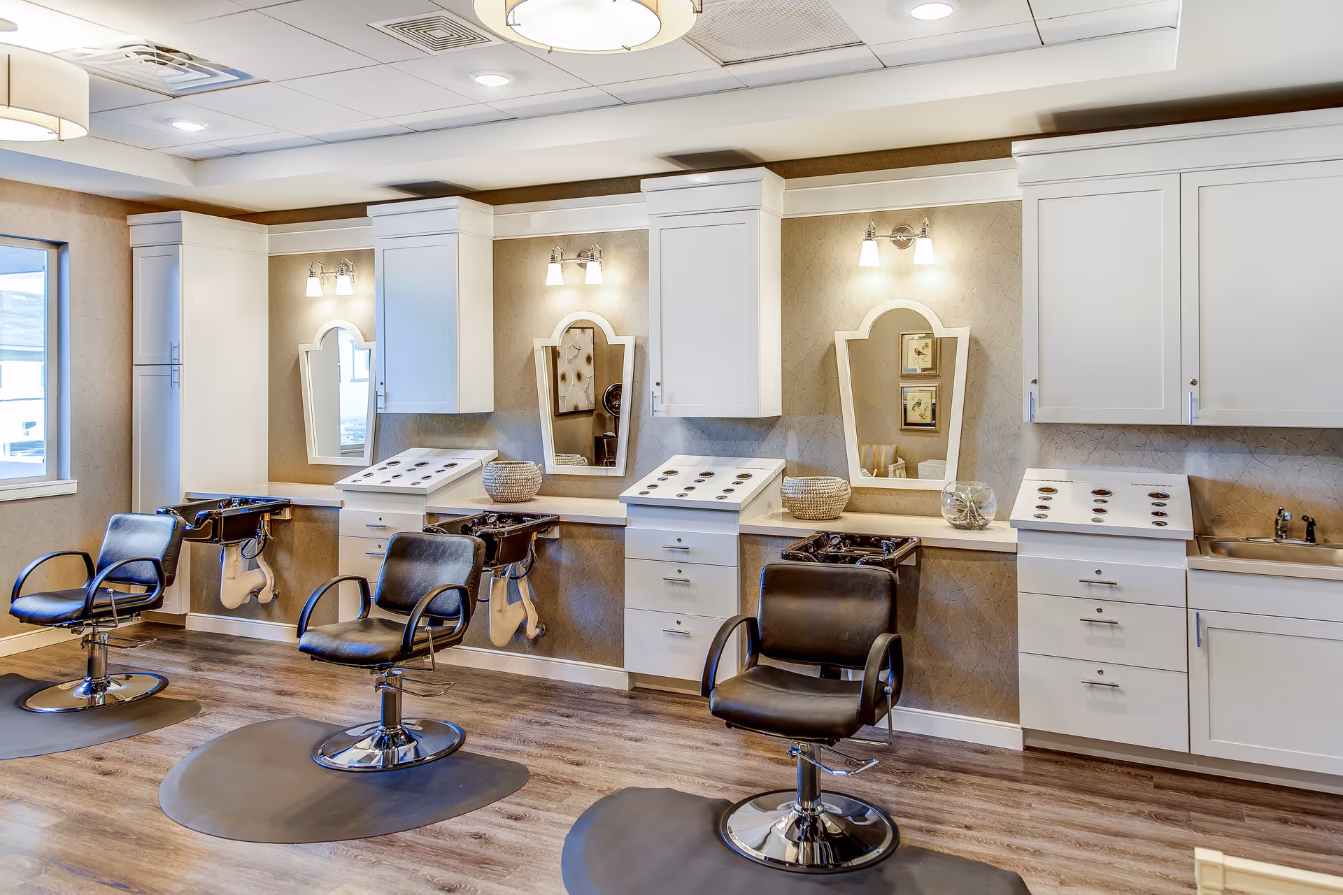 Interior view of a salon area with three black salon chairs in front of white cabinets and mirrors mounted on a beige textured wall. Each station has a small sink and hair washing basin, with overhead lights illuminating the space. The floor is wood with dark mats under each chair.