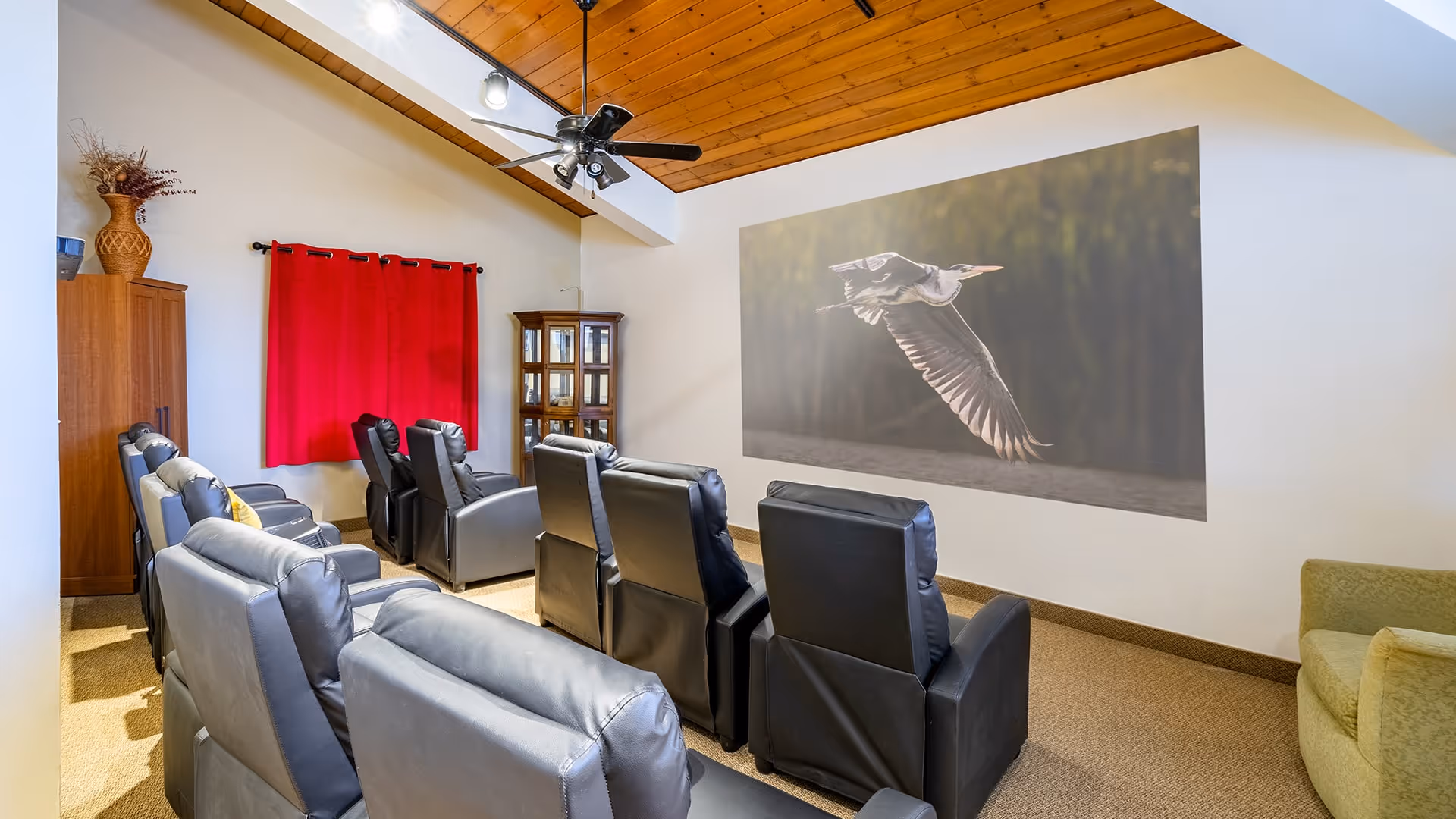 A cozy room with multiple black leather recliner chairs arranged in rows facing a large wall projection of a flying bird. The room has a wooden ceiling with a ceiling fan, a red curtain on the window, a wooden cabinet with glass doors, and a green armchair on the right side.