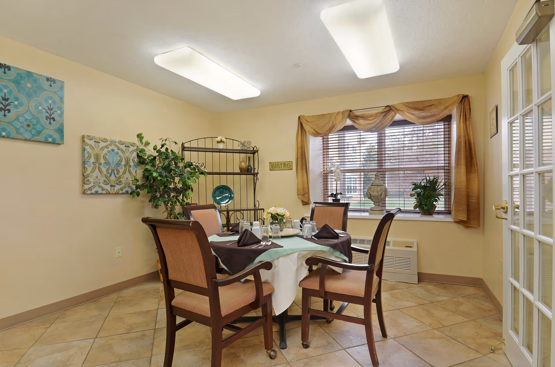 A small dining room with a round table set for four people, featuring a white tablecloth with a green overlay and dark napkins. Four wooden chairs with cushioned seats surround the table. The room has beige walls, tiled floor, a window with brown curtains, and decorative plants and artwork on the walls. A metal shelving unit is in the corner with decorative items.