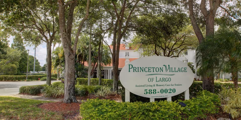 Outdoor view of Princeton Village of Largo sign surrounded by trees and shrubs with a building partially visible in the background.