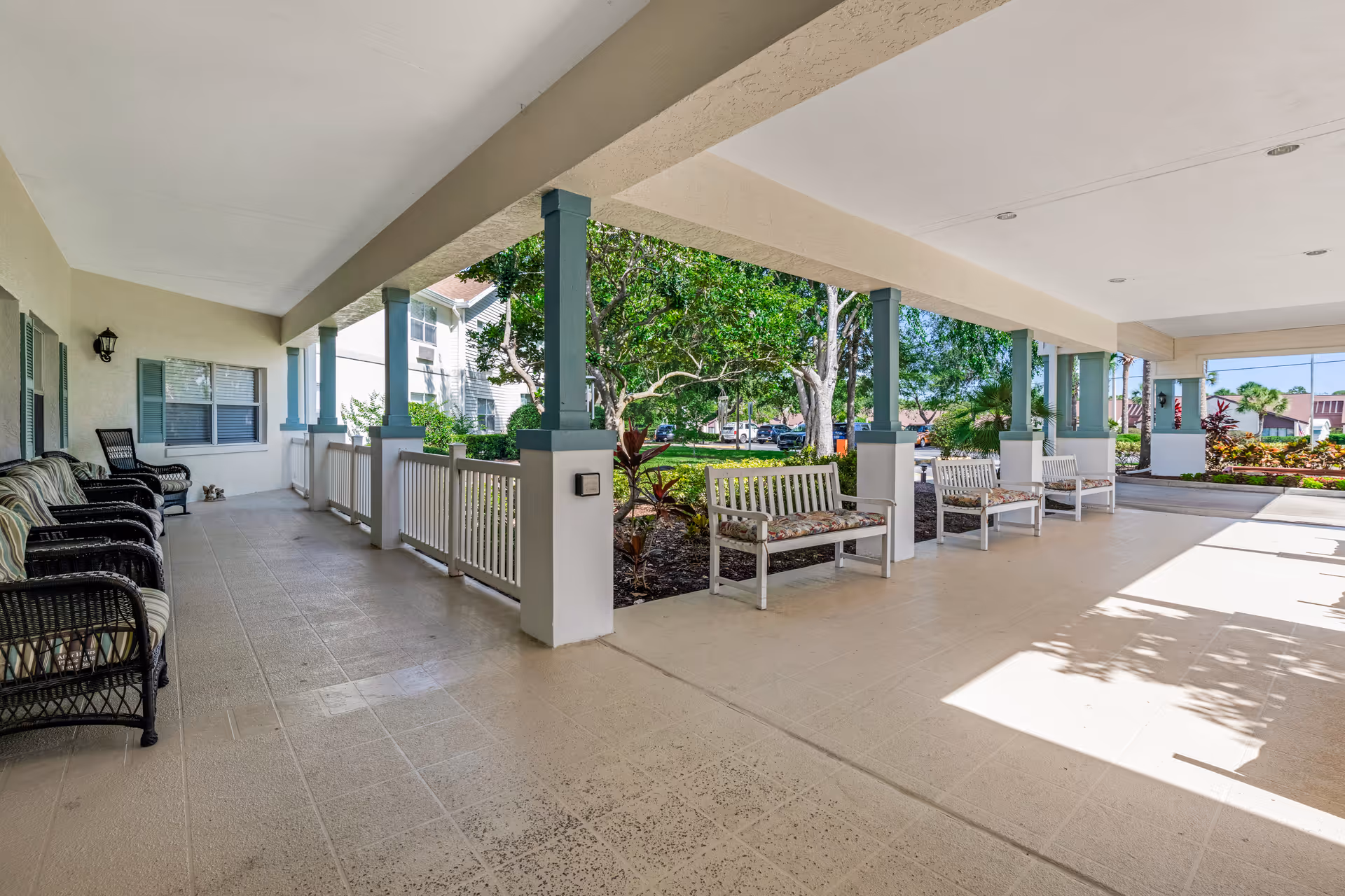 Covered outdoor patio area with multiple benches and chairs arranged along the sides. The patio overlooks a landscaped garden with trees and shrubs, and there are residential buildings visible in the background under a clear sky.