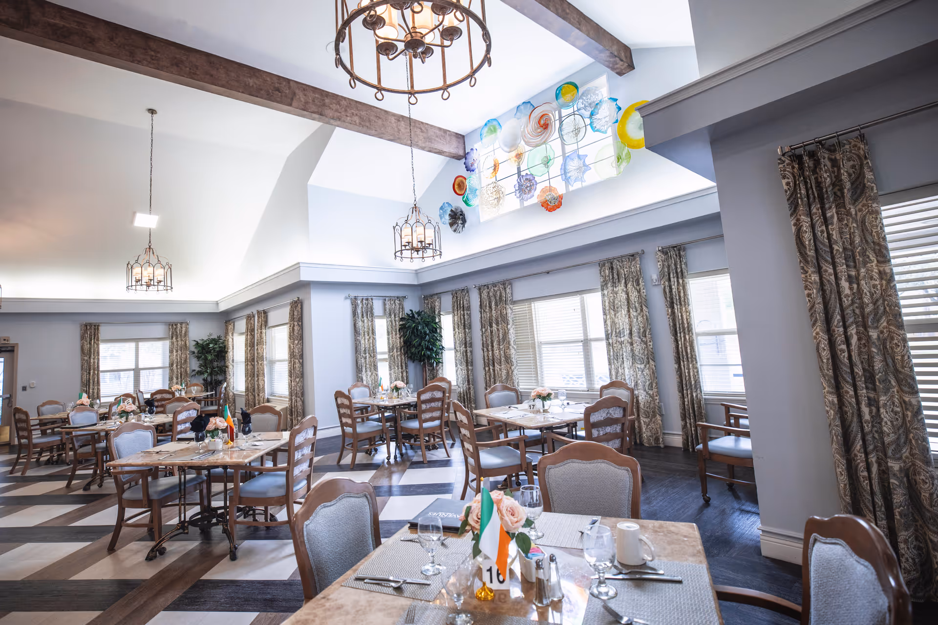 A bright and spacious dining room with multiple tables and chairs arranged neatly. Each table is set with placemats, glassware, silverware, and small floral centerpieces. The room features large windows with patterned curtains, wooden ceiling beams, and decorative hanging light fixtures. A colorful glass art installation is mounted high on one wall near the ceiling.