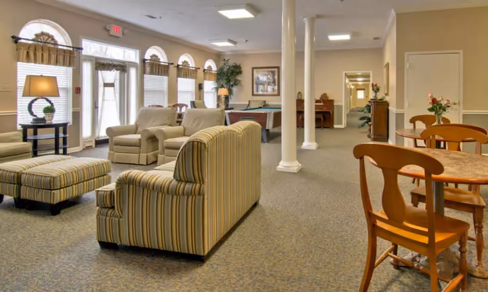 A spacious senior living facility common area with striped and beige upholstered armchairs and ottomans, wooden tables and chairs with flower vases, a pool table, and large windows with curtains allowing natural light. The room has beige walls, carpeted floors, white columns, and a hallway leading to other rooms.