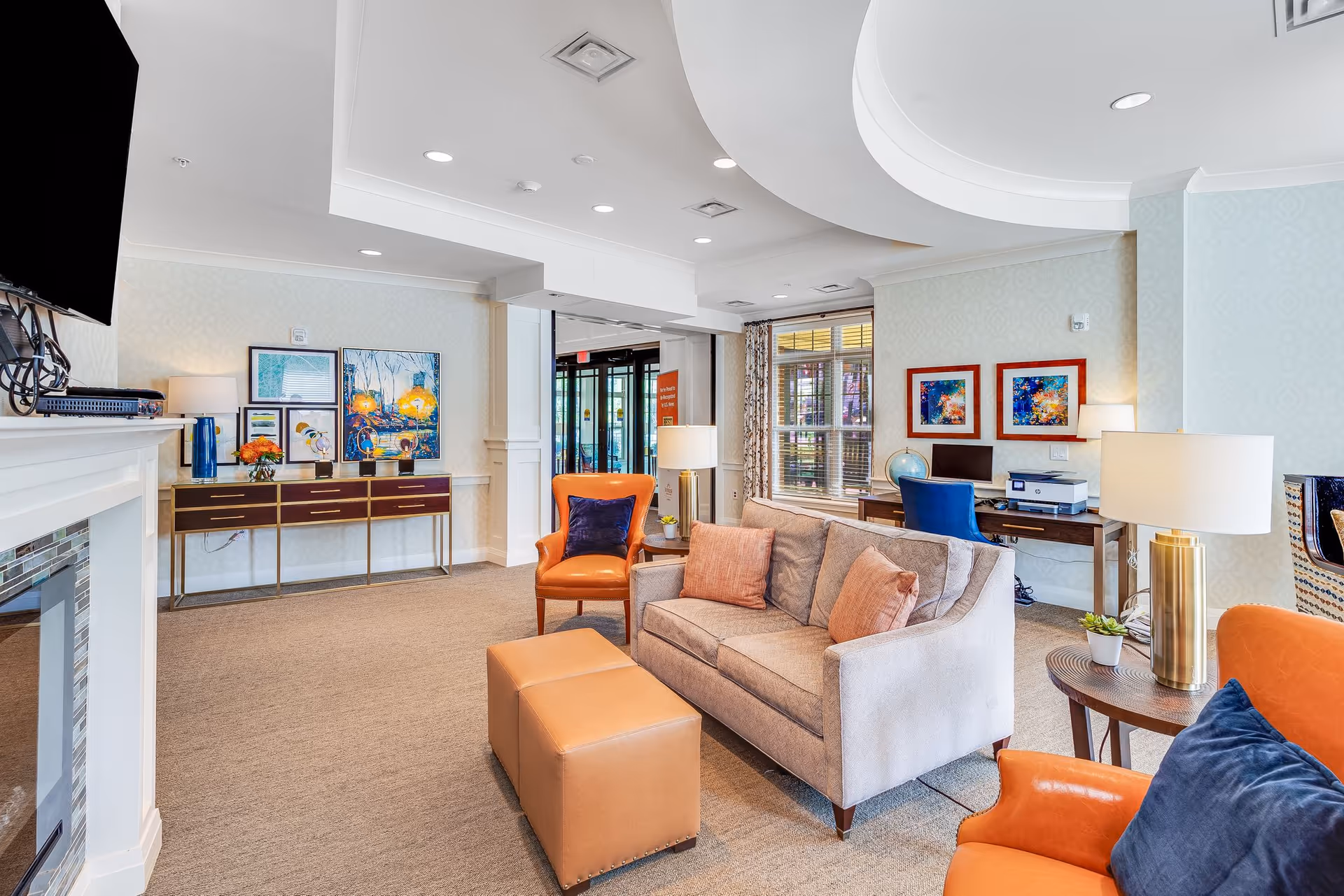 Sunny communal lounge with a sofa, orange armchairs, an ottoman, a desk area with a computer, and wall art.