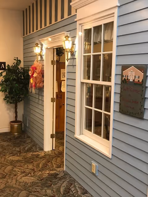 Interior hallway with blue siding walls, a white-framed window, and a door with two brass wall lanterns on either side. A welcome sign for Seneca Lake Terrace is mounted on the wall next to the window. There is a potted plant on the left side and patterned carpet on the floor.