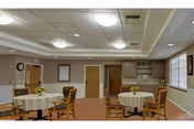 A well-lit common area with round tables covered with white tablecloths and wooden chairs arranged around them. The room has a beige carpet, light-colored walls, a ceiling with recessed lighting, and a small kitchenette area with cabinets and a microwave. There are windows on the right side letting in natural light and a clock on the wall to the left.