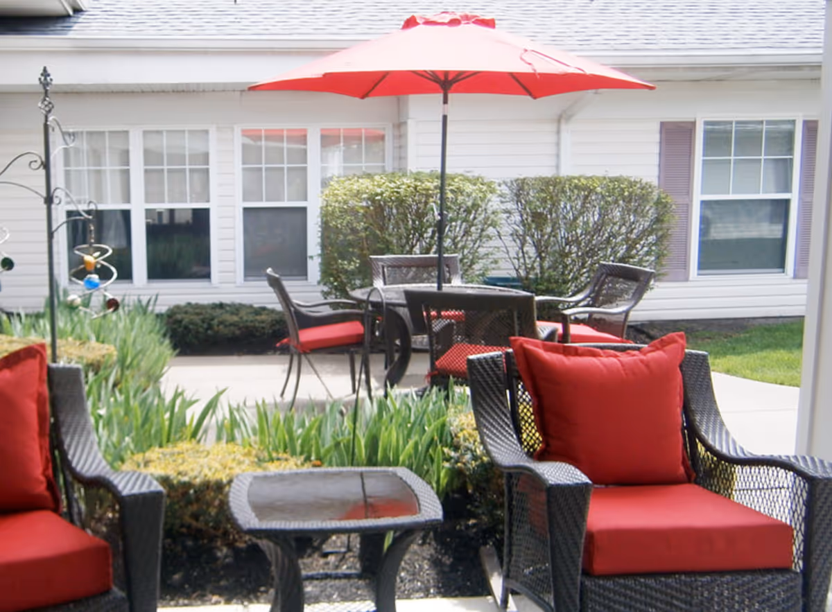 Outdoor patio area with black wicker chairs and red cushions, a small glass-top table, and a round table with matching chairs under a red umbrella. The patio is surrounded by green plants and bushes, with a white building with windows in the background.