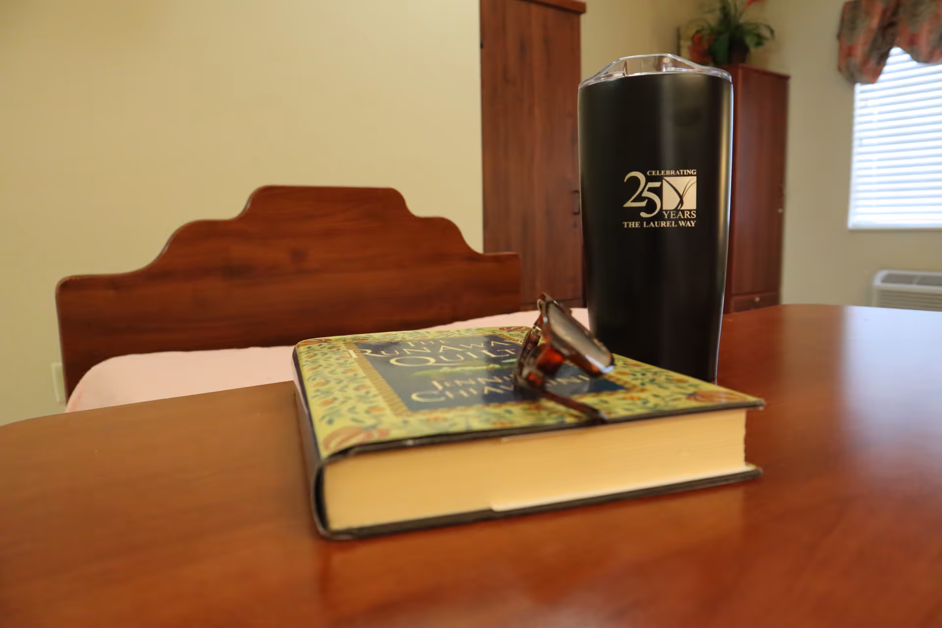 A wooden table with a closed book, a pair of eyeglasses resting on the book, and a black tumbler with the text 'Celebrating 25 Years The Laurel Way'. In the background, there is a wooden headboard, a cabinet, a window with blinds, and a potted plant on top of the cabinet.
