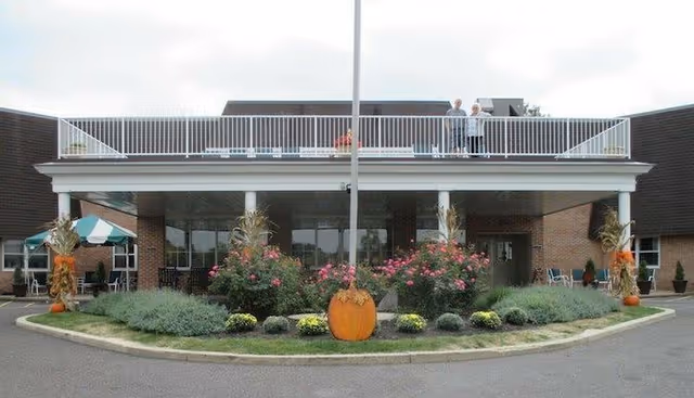 Front exterior view of Manatawny Manor Center for Rehabilitation and Nursing building with a covered entrance, landscaped garden featuring pumpkins and flowers, and two people standing on the rooftop balcony.