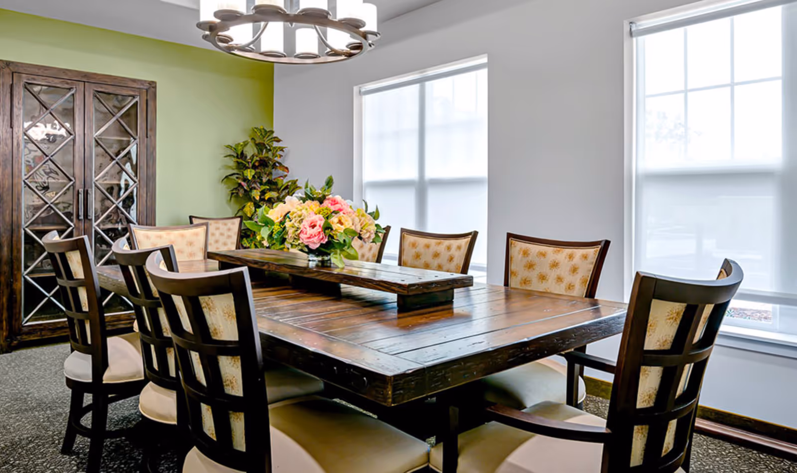 A dining room with a large wooden table surrounded by eight cushioned chairs with patterned backs. A floral centerpiece is placed on the table. The room has two large windows with white blinds, a green accent wall, a wooden cabinet with glass doors, and a modern circular chandelier hanging from the ceiling.