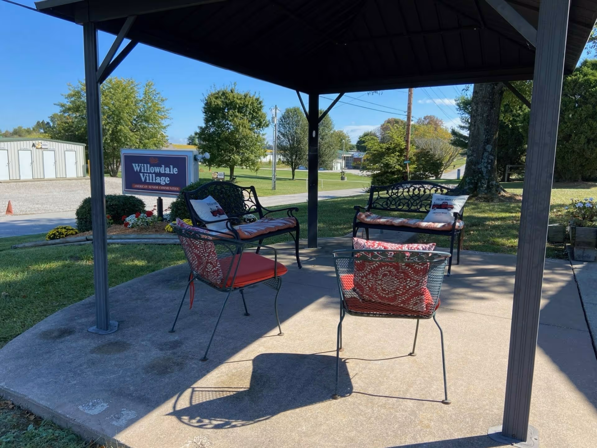 Covered outdoor seating under a gazebo with metal chairs and benches on a concrete pad and a 'Willowdale Village' sign in the background.