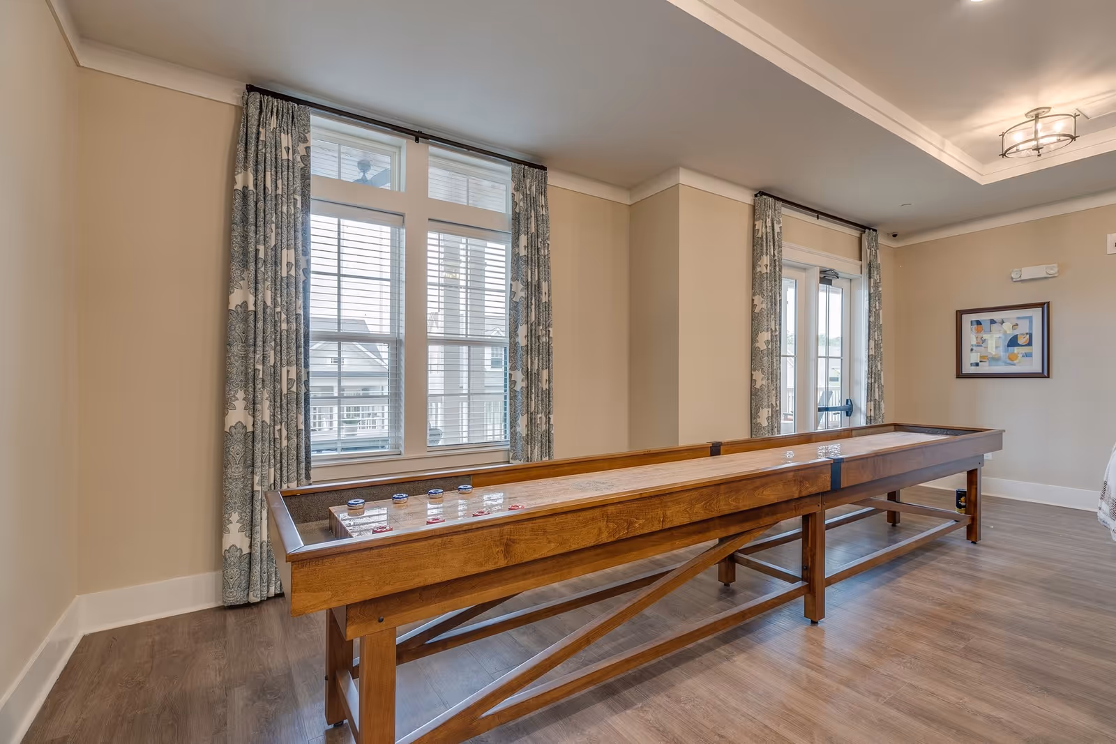 A spacious room with a wooden shuffleboard table in the center, large windows with patterned curtains allowing natural light to enter, light beige walls, wood flooring, and a framed abstract artwork on the wall.