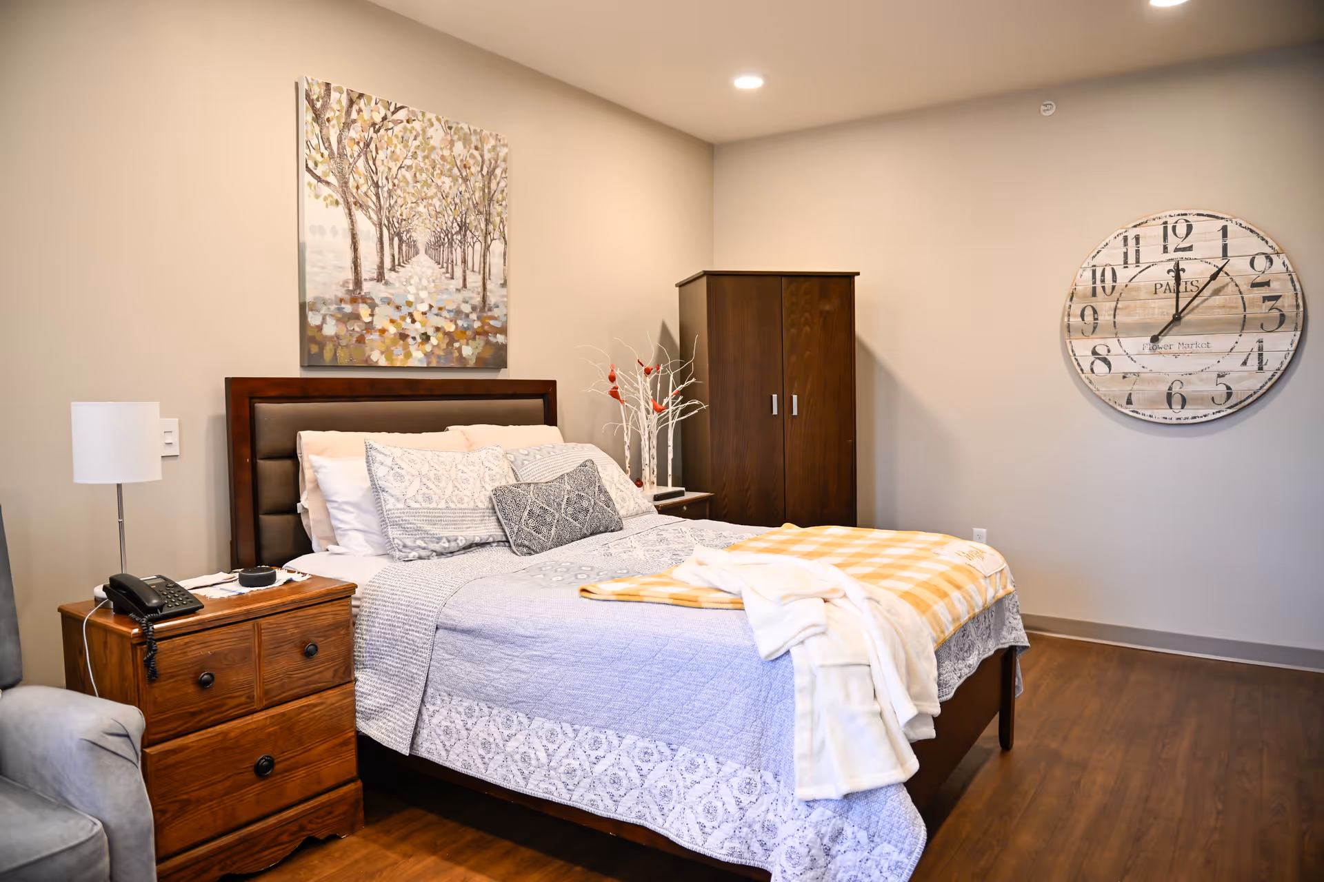 A cozy bedroom with a wooden bed frame, neatly made bed with patterned bedding and a yellow checkered blanket. There is a wooden nightstand with a lamp and a telephone, a wooden wardrobe, a large round wall clock, and a painting of a tree-lined path on the wall. The room has wooden flooring and soft lighting.