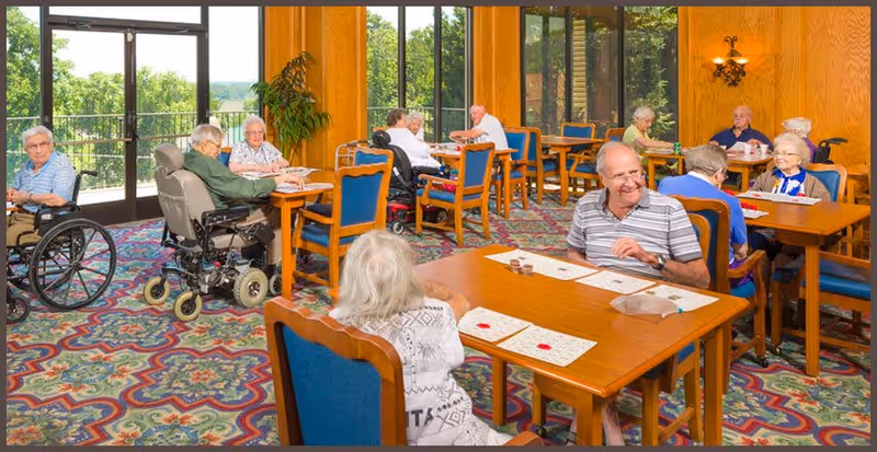 A group of elderly people sitting at wooden tables in a brightly lit room with large windows and colorful patterned carpet. Some individuals are in wheelchairs, and they appear to be engaged in playing bingo or a similar game, with bingo cards and markers on the tables.